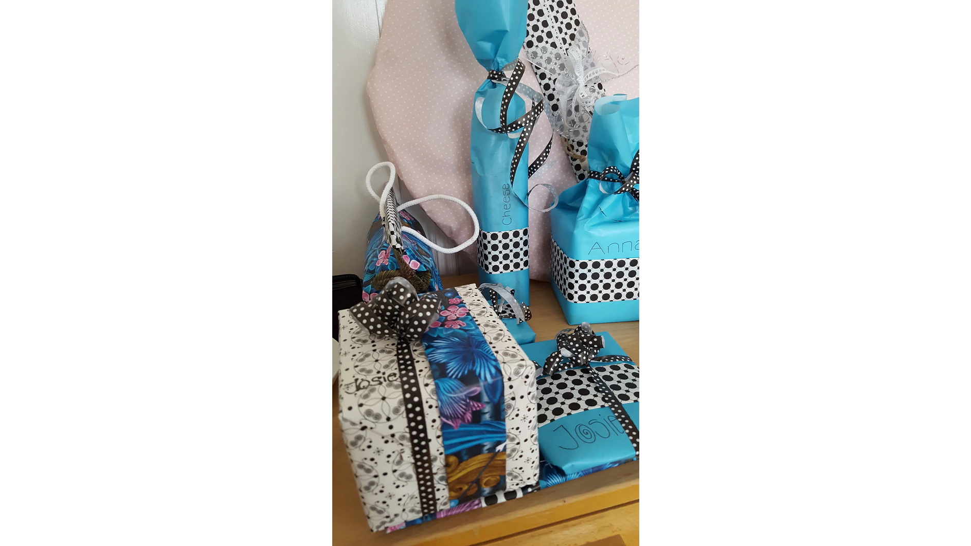 Auf einem Tisch liegen mehrere verpackte Geschenke. Die Pakete sind mit blauen und floralen Mustern und schwarz-weißen gepunkteten Bändern versehen. Jedes Geschenk hat ein Namensschild, das mit Tesa-Klebeband befestigt ist, und der Hintergrund besteht aus rosa gepunktetem Stoff. (This text has been generated by AI)