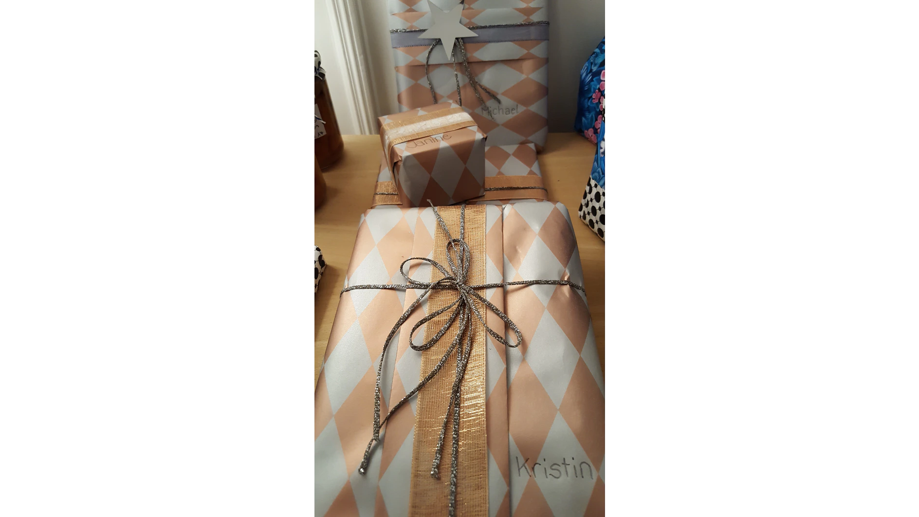 Eine Gruppe hübsch verpackter Geschenke in metallischem Rautenpapier mit silbernen Kordeln und einer goldenen Schleife, alles mit Tesa-Klebeband befestigt. Auf einigen der Pakete sind Namen geschrieben, und sie liegen auf einer Holzoberfläche, neben anderen verpackten Geschenken. (This text has been generated by AI)