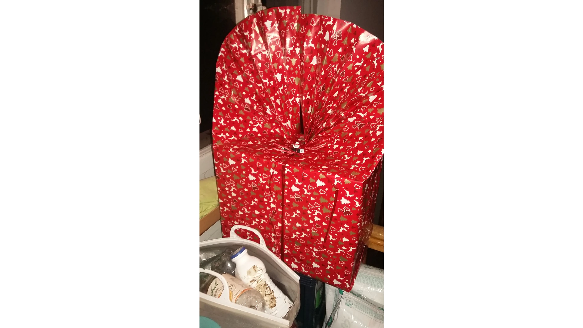 Ein großes Geschenk, eingewickelt in rotes Papier mit weißen Feiertagsmustern und einer spektakulären fächerartigen Oberseite. Das Geschenk wird mit Tesa-Klebeband befestigt und auf einer Oberfläche in der Nähe eines Fensters platziert. Daneben steht eine Tüte mit verschiedenen Haushaltsgegenständen. (This text has been generated by AI)