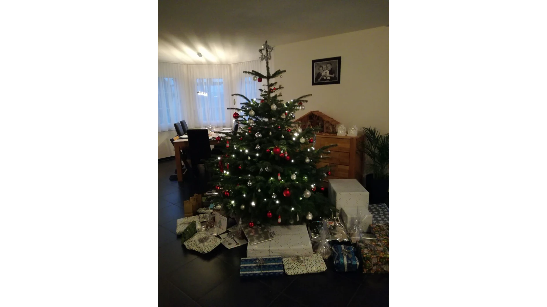 In einem Raum steht ein geschmückter Weihnachtsbaum mit roten und silbernen Ornamenten und einem Stern auf der Spitze. Darunter stapeln sich verpackte Geschenke, die sorgfältig mit Tesaband befestigt sind. Im Hintergrund sind ein Esstisch mit Stühlen und ein Holzschrank mit einer Weihnachtskrippe zu sehen. (This text has been generated by AI)