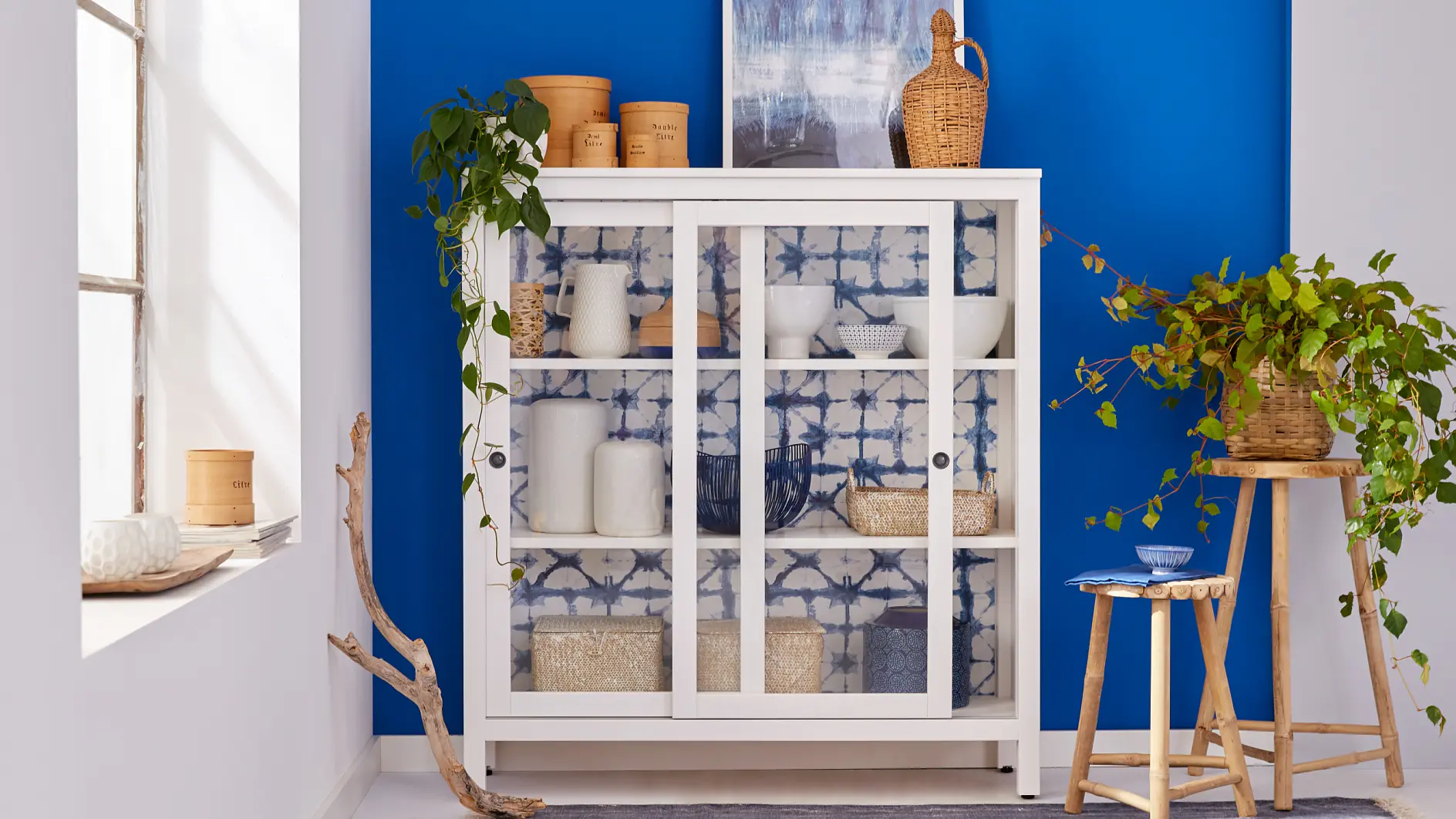 A white cabinet with glass doors stands against a bright blue wall. Inside, various white and wicker items are displayed. A wooden stool with a plant and bowl is beside the cabinet. A leafy branch decorates the area next to a large window.