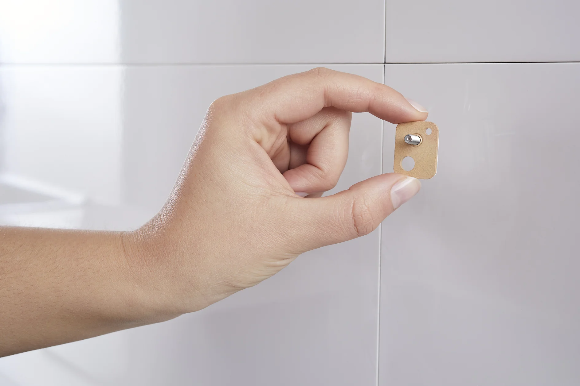 A hand applying a small square adhesive hook with a metal peg onto a white tiled wall using tesa tape. (This text has been generated by AI)