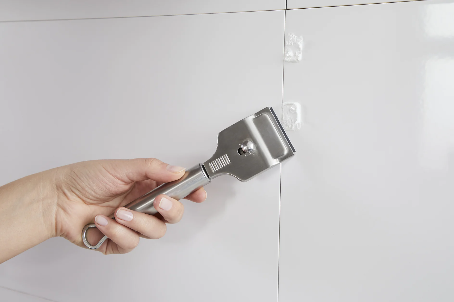 A hand is holding a metal scraper against a white tiled wall. There are small, hardened white blobs on the wall being scraped off. The scraper has a shiny metal blade and a textured handle. The tiles have visible grout lines, and strips of tesa tape can be seen along some of the edges, ensuring neatness in any sticky situation. (This text has been generated by AI)