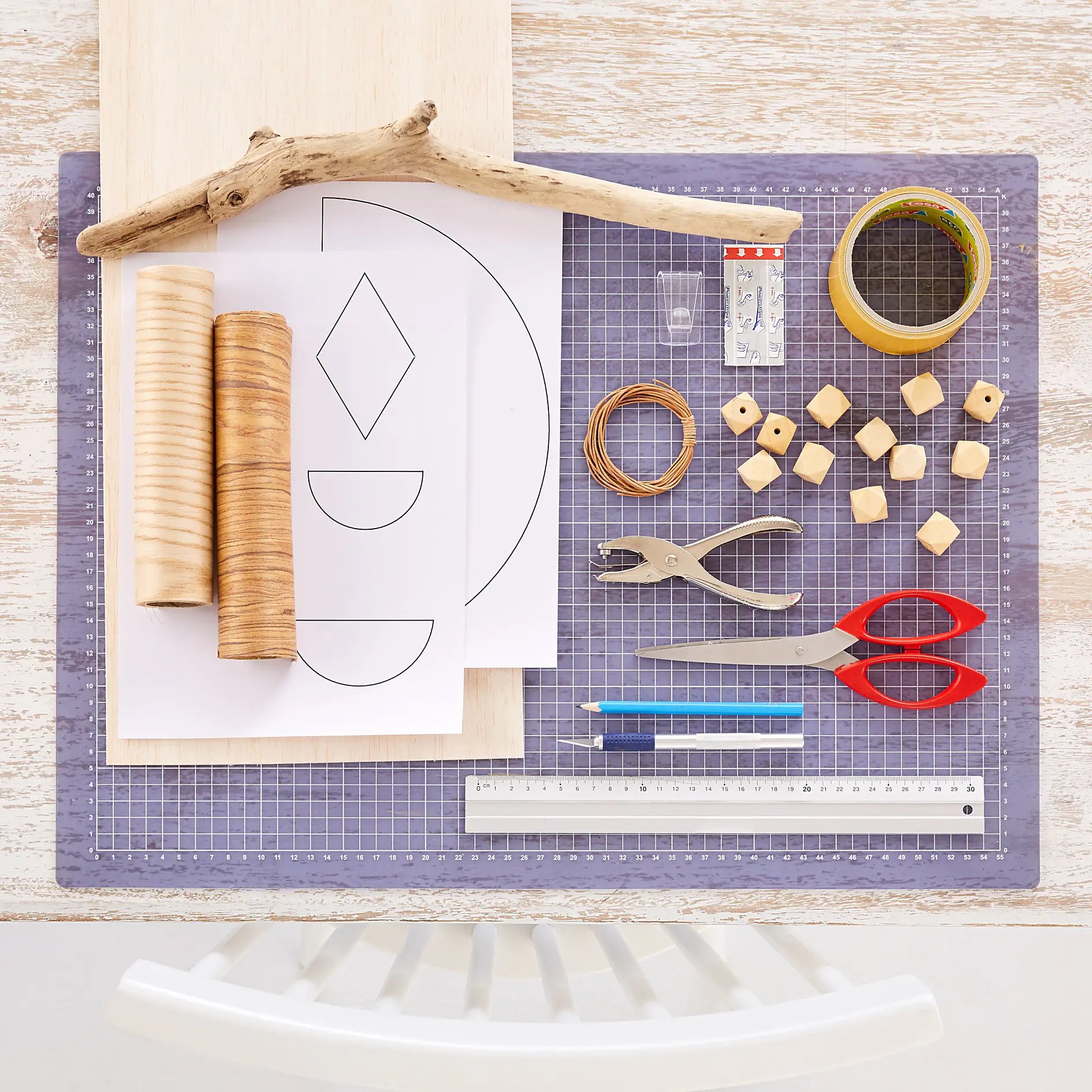 A crafting workspace featuring a cutting mat with various items: geometric pattern papers, a wooden board, a branch, wooden cylinders and cubes, a ruler, scissors, hole punch, pencils, tesa tape adhesive tape, and wire. (This text has been generated by AI)