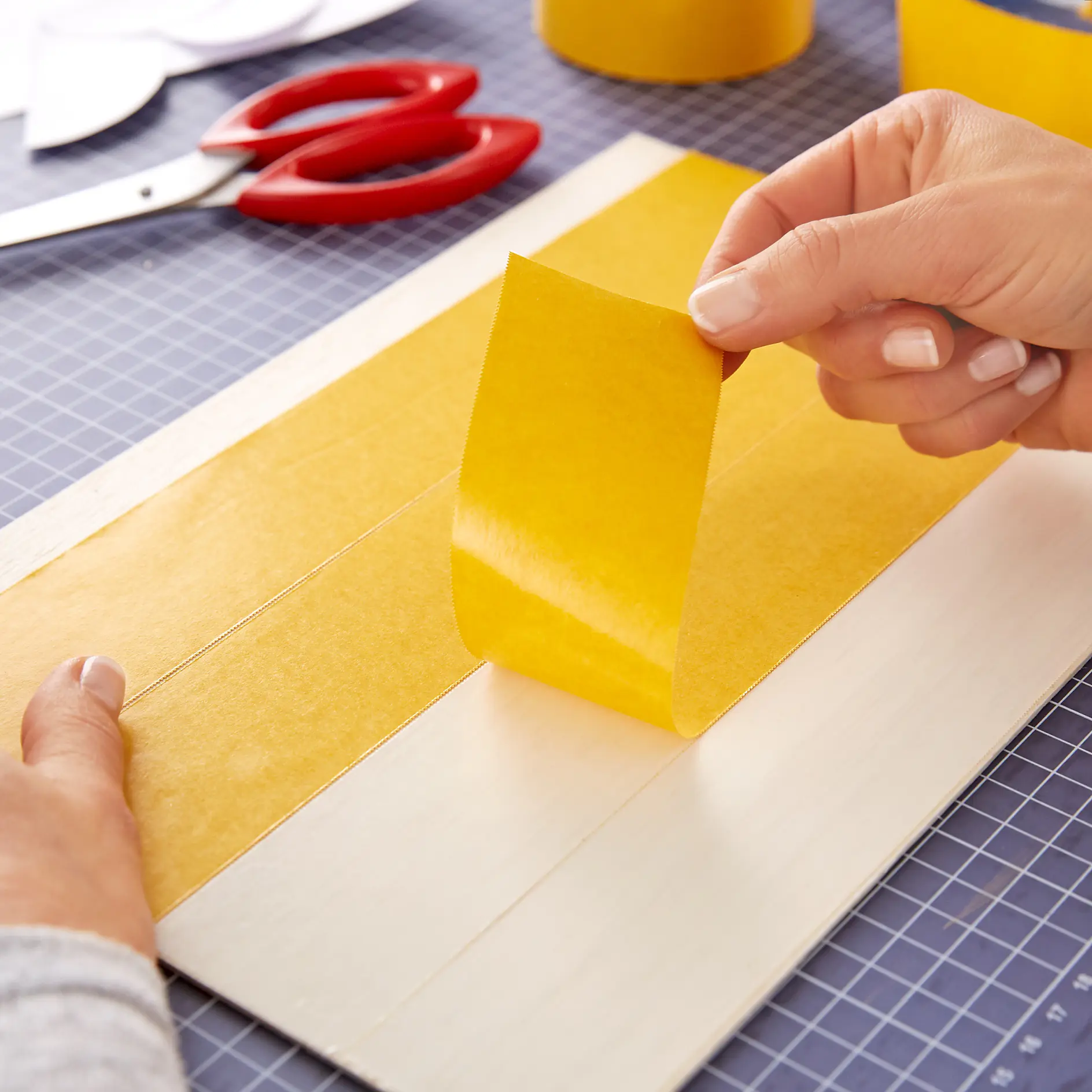 Hands peeling yellow tesa tape from a sheet on a cutting mat. Scissors and more tesa tape can be seen in the background. (This text has been generated by AI)