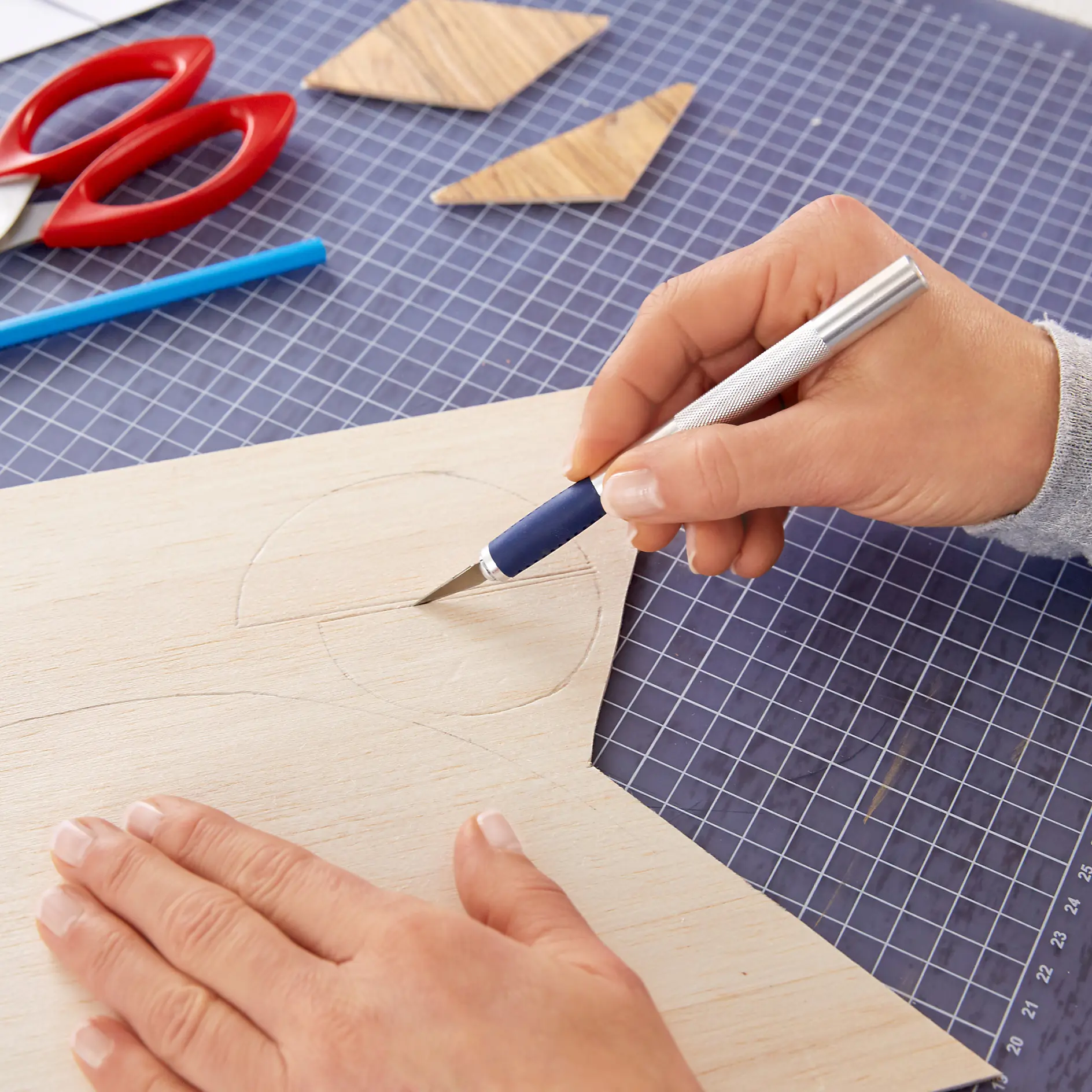 A person uses a craft knife to cut a round shape out of a piece of plywood on a grid cutting mat. Red scissors, a blue pencil, and two wooden triangle cutouts are nearby. To assist with measuring and holding the plywood in place during cutting, pieces of tesa tape are used effectively around the workspace. (This text has been generated by AI)