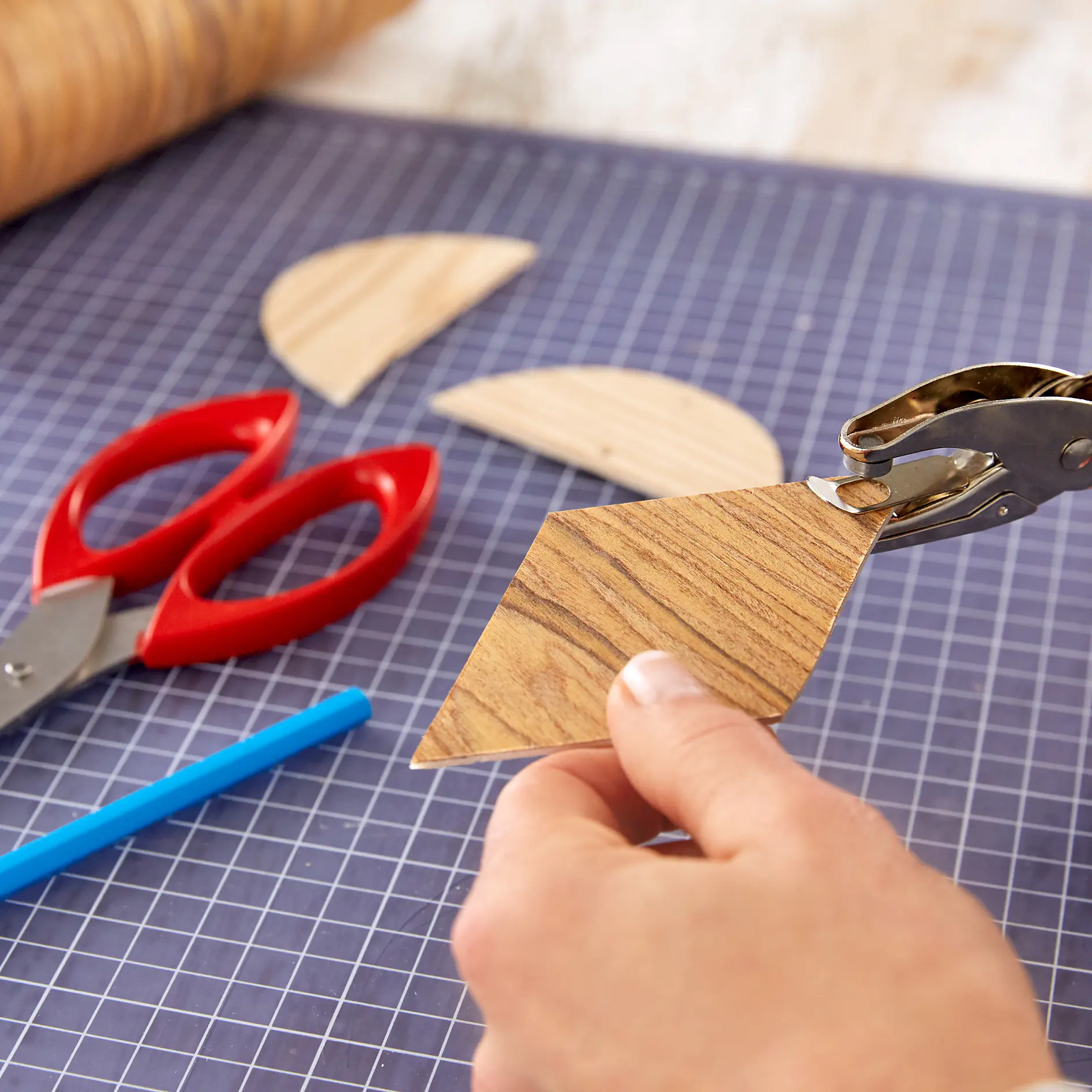 A hand uses a hole puncher on a piece of wood-patterned paper atop a grid-patterned surface. Red scissors and a blue pen lie nearby, along with additional paper pieces. (This text has been generated by AI)