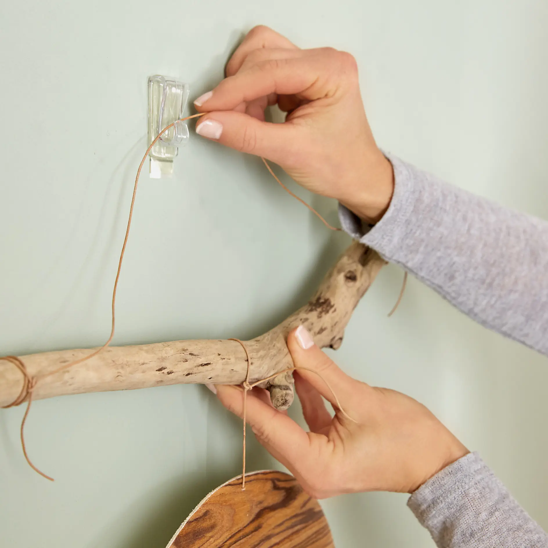 Hands attaching a piece of driftwood to a wall with brown string and a clear adhesive hook. The person wears a long-sleeve gray shirt, and the wall is light green. (This text has been generated by AI)