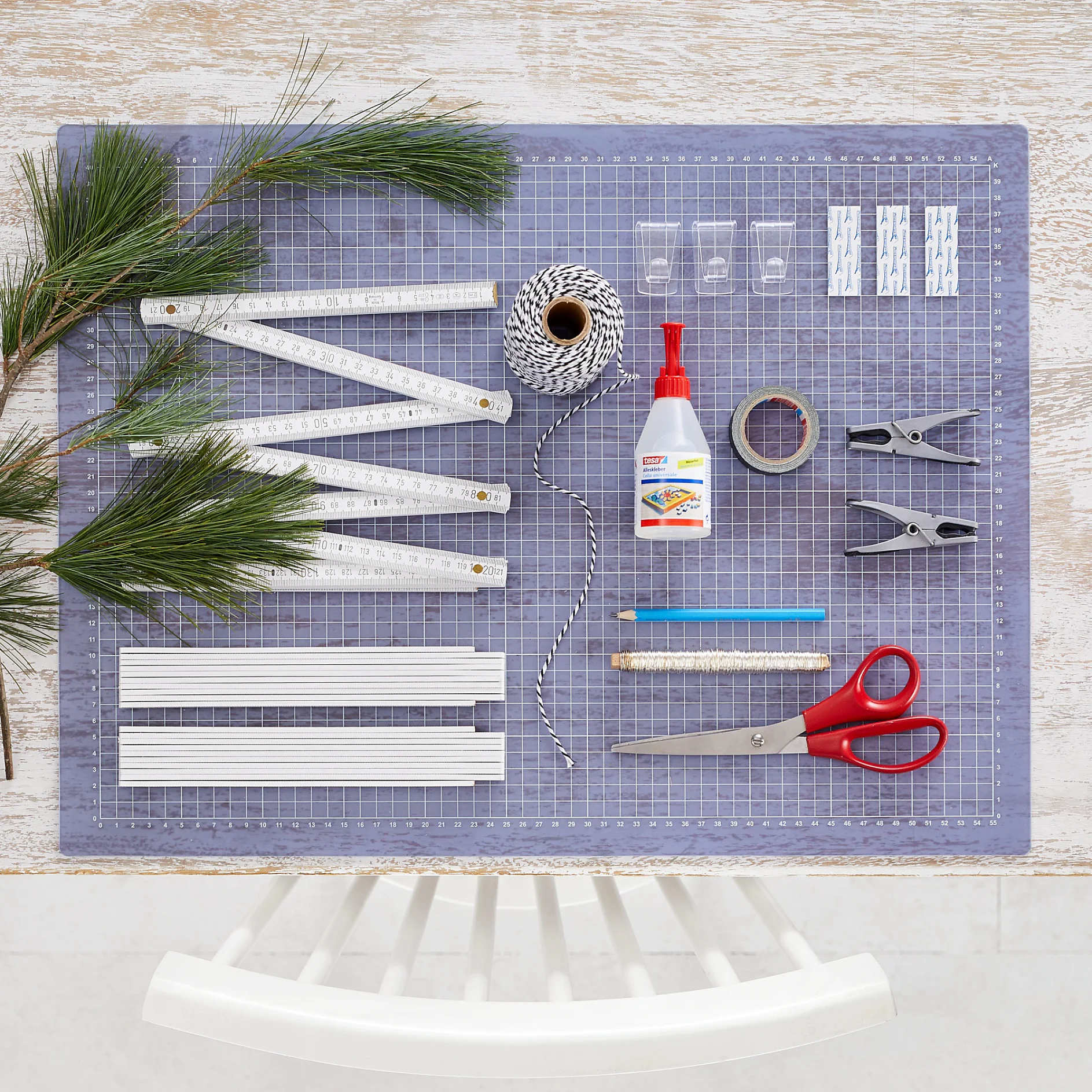 A top view of a crafting table displaying various items: pine branches, a folding ruler, string, glue, tesa tape, clothespins, clips, a pencil, scissors, and plastic binders. The table has a gridded cutting mat underneath the items. (This text has been generated by AI)