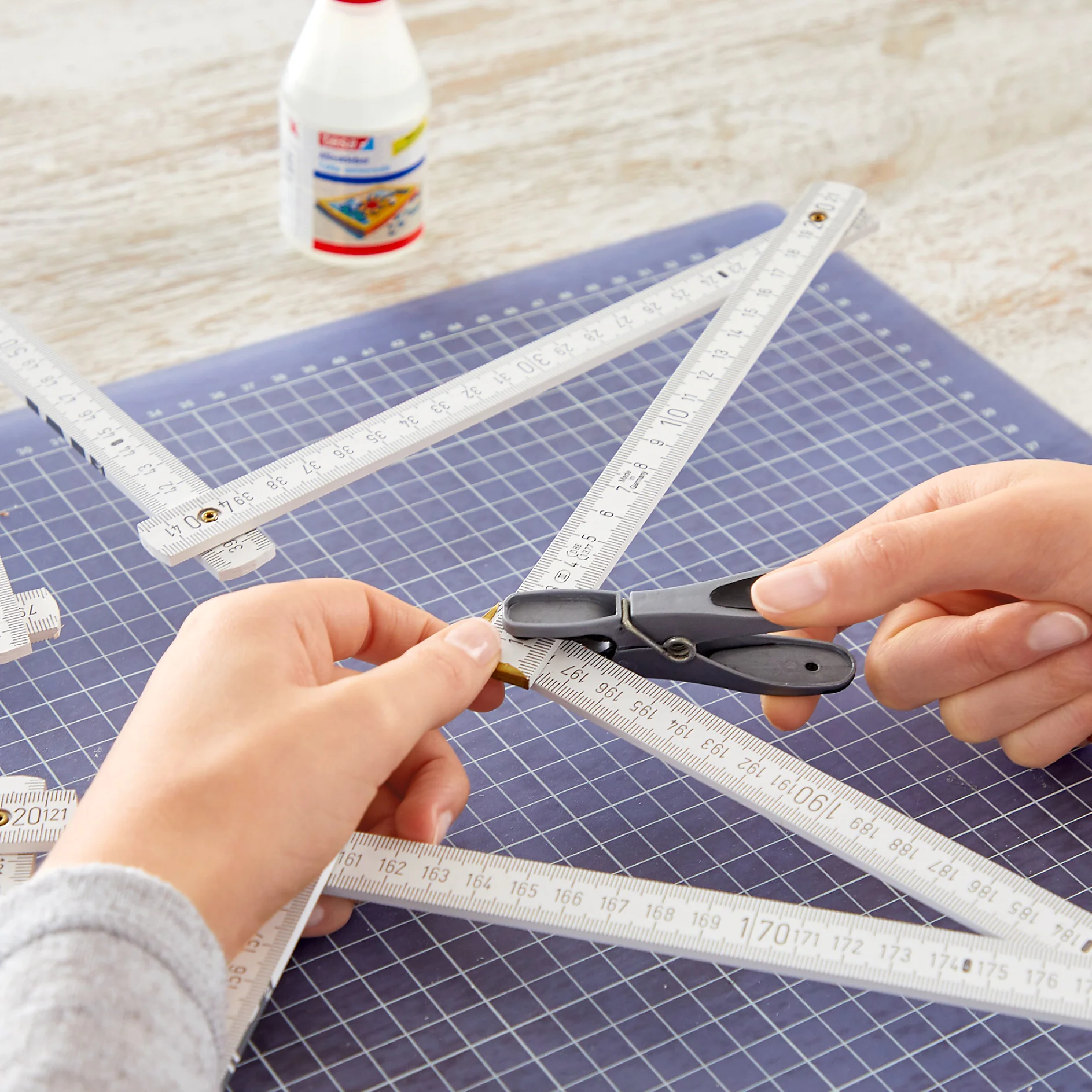 Two hands use scissors to cut an object while a white folding ruler measures on a blue grid mat. A bottle of tesa tape adhesive is visible in the background on a wooden surface. (This text has been generated by AI)