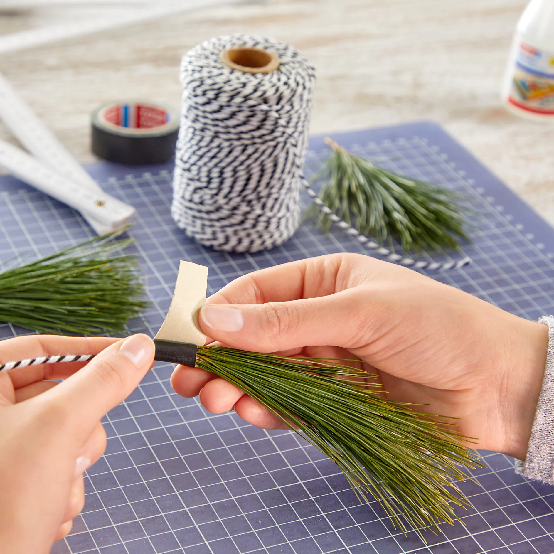 Hands are seen tying a bundle of pine needles with black and white twine. A strip of tesa tape is being wrapped around the base. The background shows more pine bundles, a spool of twine, tesa tape, a ruler, and a grid-patterned mat. (This text has been generated by AI)