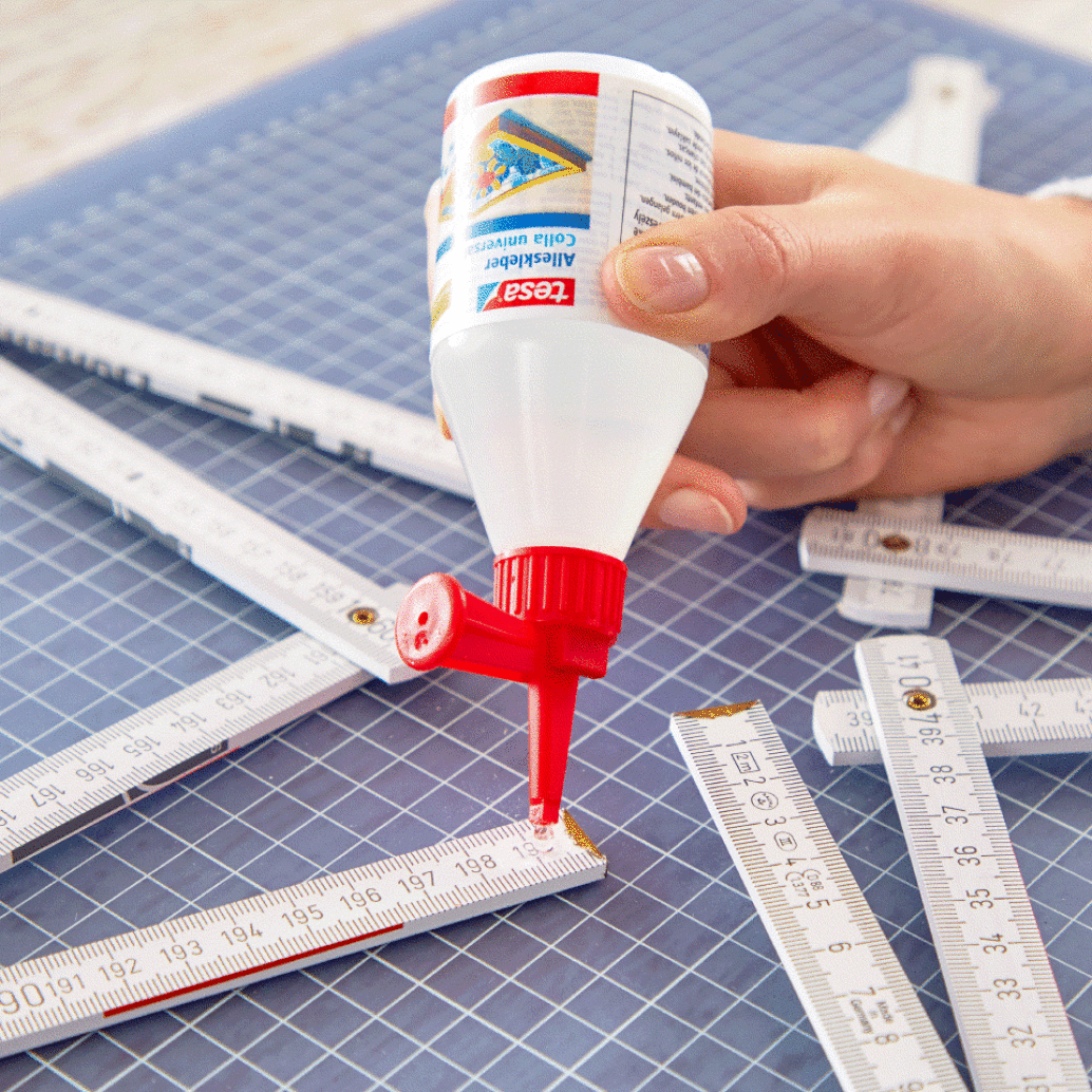 A person applies tesa tape to a folding ruler on a cutting mat. The ruler is partly unfolded and lies alongside similar tools. The glue bottle has a red cap and is being held in the right hand. The background mat is blue with a grid pattern. (This text has been generated by AI)