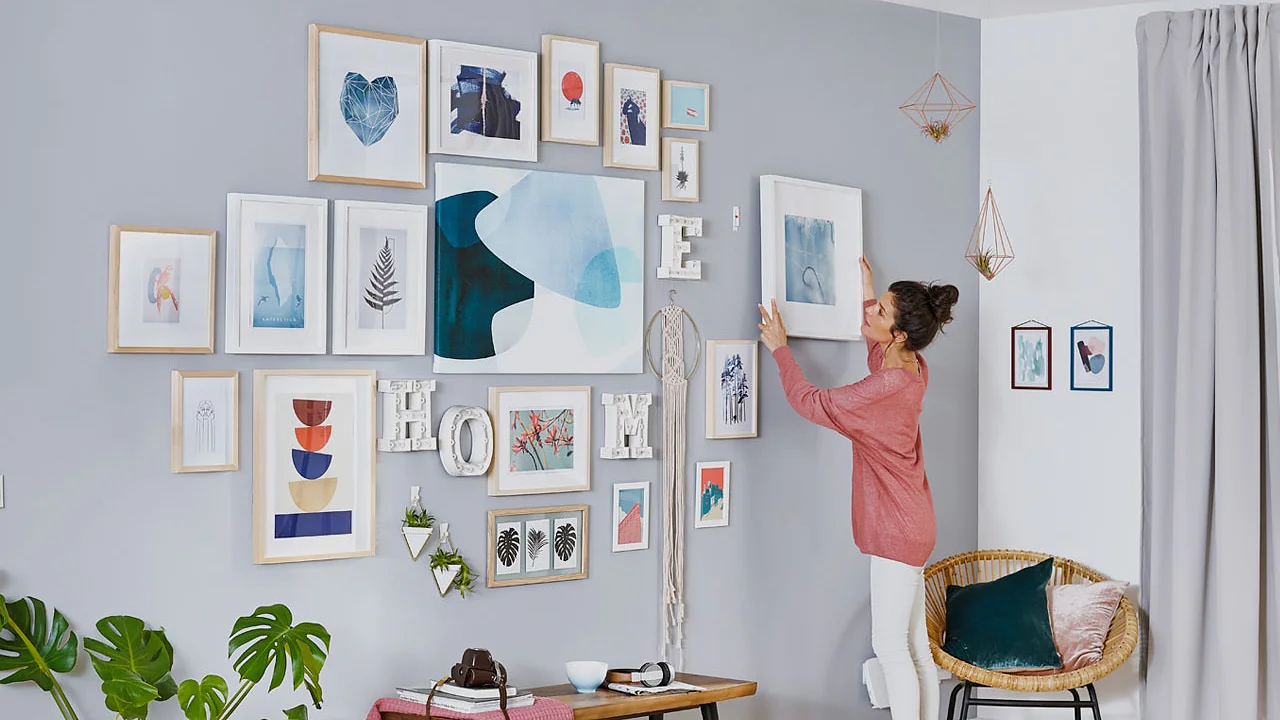 A woman in a pink shirt is arranging frames on a grey and white wall, filled with various artwork and photos, using tesa tape. The room has a modern design with a small kitchen on the left, light wood flooring, and green plants for decoration. (This text has been generated by AI)