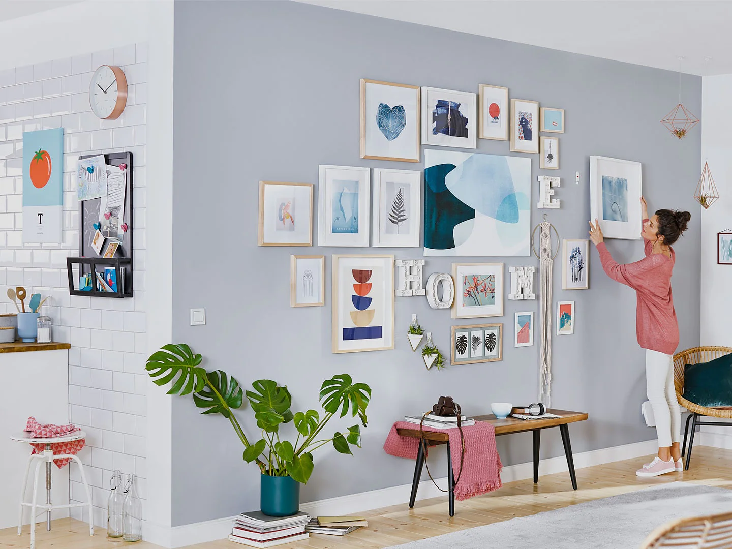 A woman in a pink shirt is arranging frames on a grey and white wall, filled with various artwork and photos, using tesa tape. The room has a modern design with a small kitchen on the left, light wood flooring, and green plants for decoration. (This text has been generated by AI)