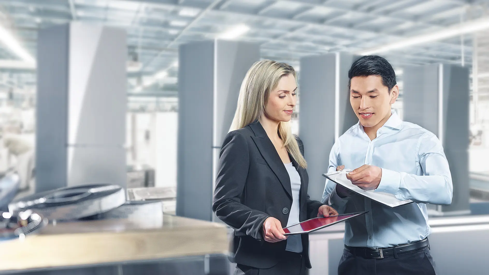 A man and a woman are discussing documents in a bright industrial setting. The woman holds a tablet, and the man holds a clipboard secured with tesa tape. They are both professionally dressed. Equipment and large metal structures are visible in the background.