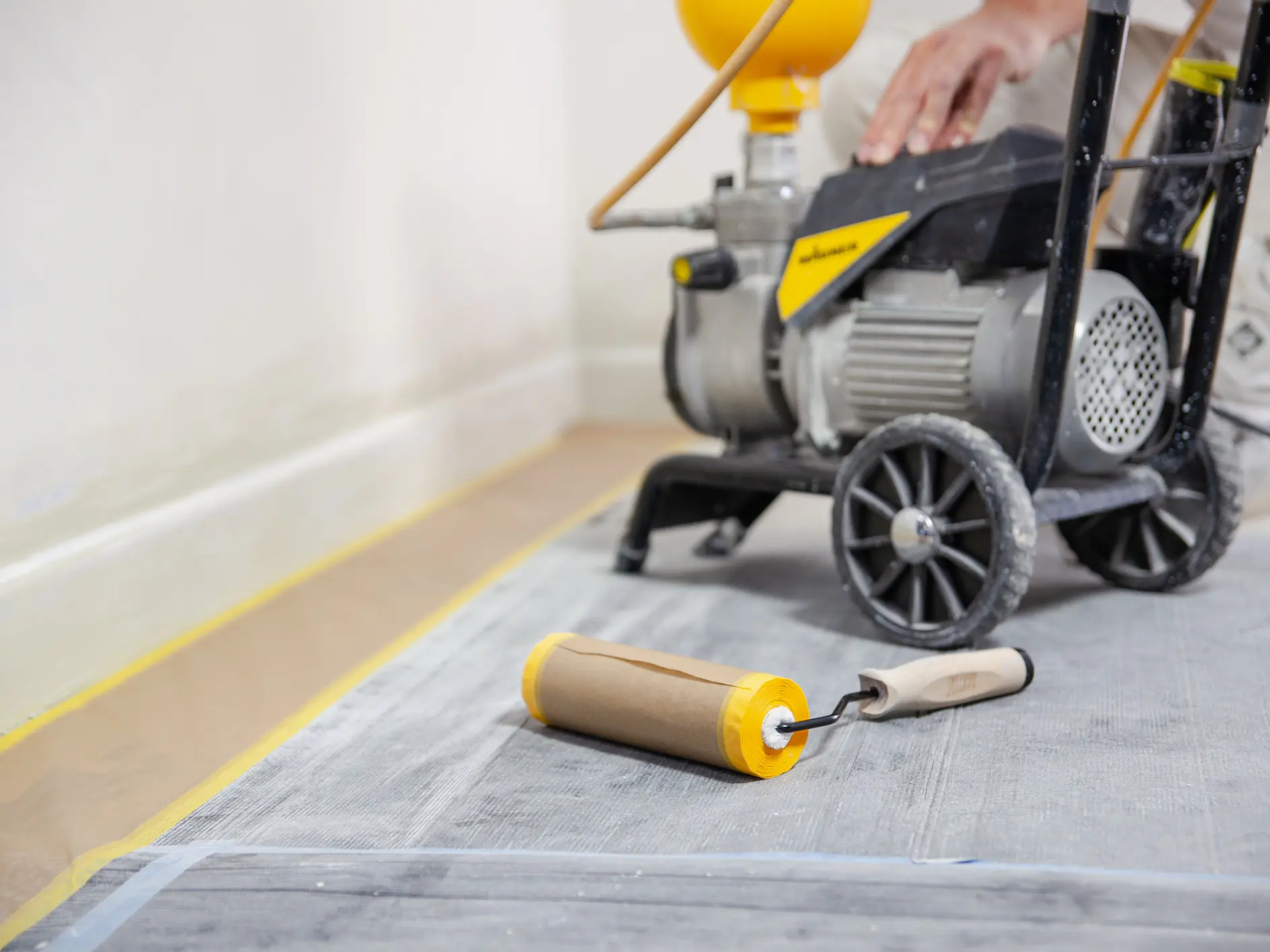 A paint roller lies on a protective floor covering. In the background, a person operates a paint spraying machine near a wall. tesa tape is used to secure various surfaces and edges, ensuring clean lines. The scene suggests a room being prepared or in the process of being painted. (This text has been generated by AI)