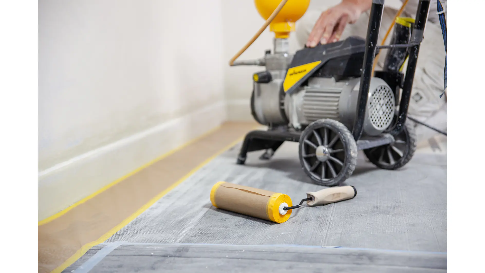 A paint roller lies on a protective floor covering. In the background, a person operates a paint spraying machine near a wall. tesa tape is used to secure various surfaces and edges, ensuring clean lines. The scene suggests a room being prepared or in the process of being painted. (This text has been generated by AI)