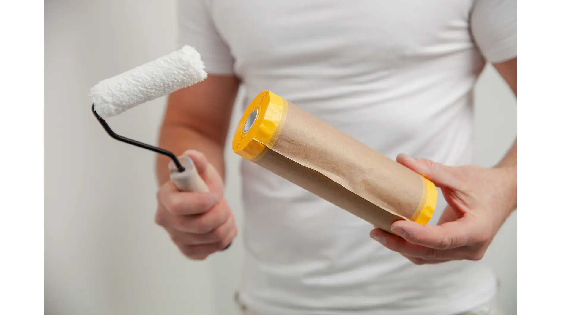 A person wearing a white shirt holds a small white paint roller in one hand and a rolled-up sheet of brown paper with yellow edges tesa tape in the other hand. The background is plain white. (This text has been generated by AI)