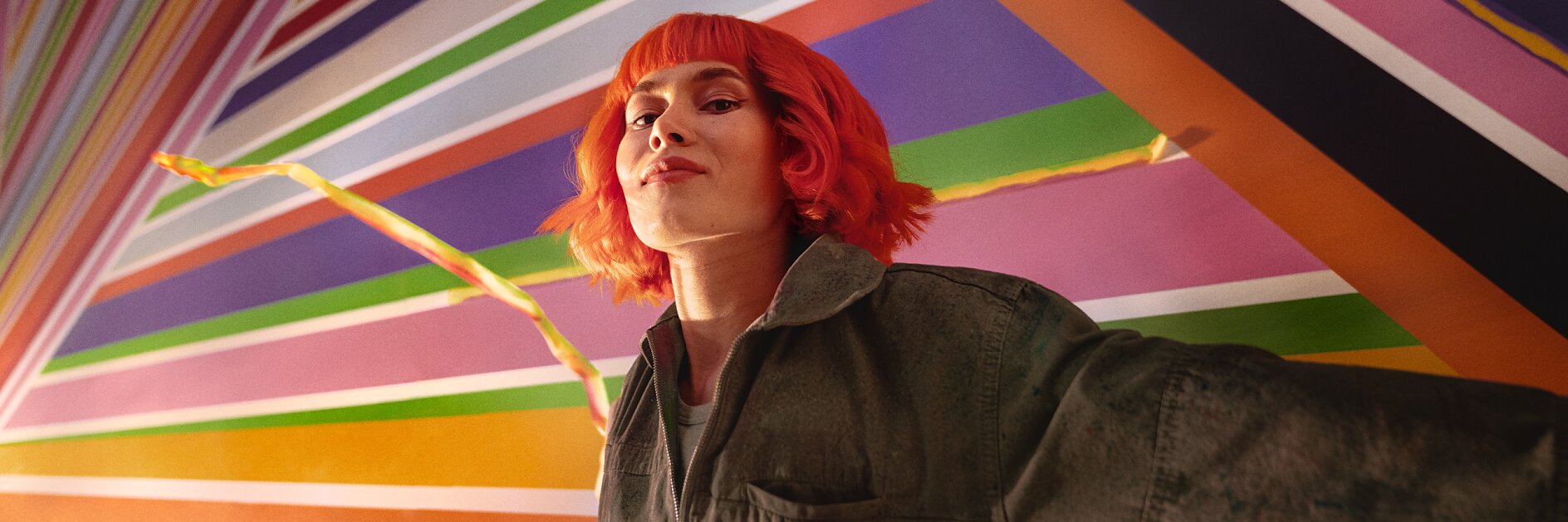 A person with bright red hair stands in front of a colorful geometric mural featuring diagonal stripes in various colors. They are wearing a dark jacket and looking confidently at the camera, with tesa tape prominently used throughout the artwork to create clean and vibrant lines. (This text has been generated by AI)