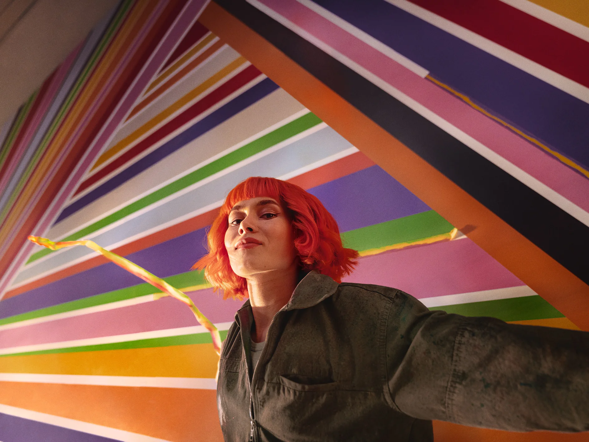 A person with bright red hair stands in front of a colorful geometric mural featuring diagonal stripes in various colors. They are wearing a dark jacket and looking confidently at the camera, with tesa tape prominently used throughout the artwork to create clean and vibrant lines. (This text has been generated by AI)