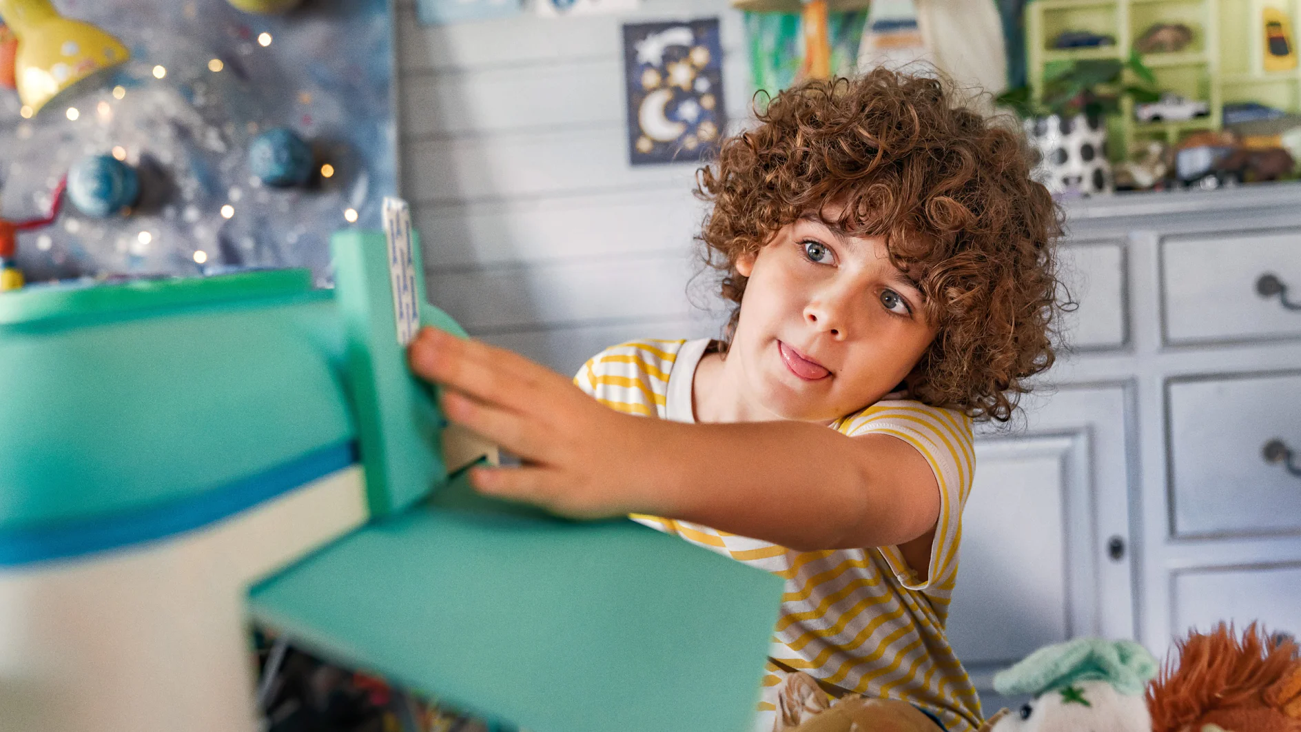 A young child with curly hair and a striped yellow shirt plays with a toy. The room has colorful decorations, a lamp, and a dresser in the background. The child appears focused and has their tongue slightly out. (This text has been generated by AI)