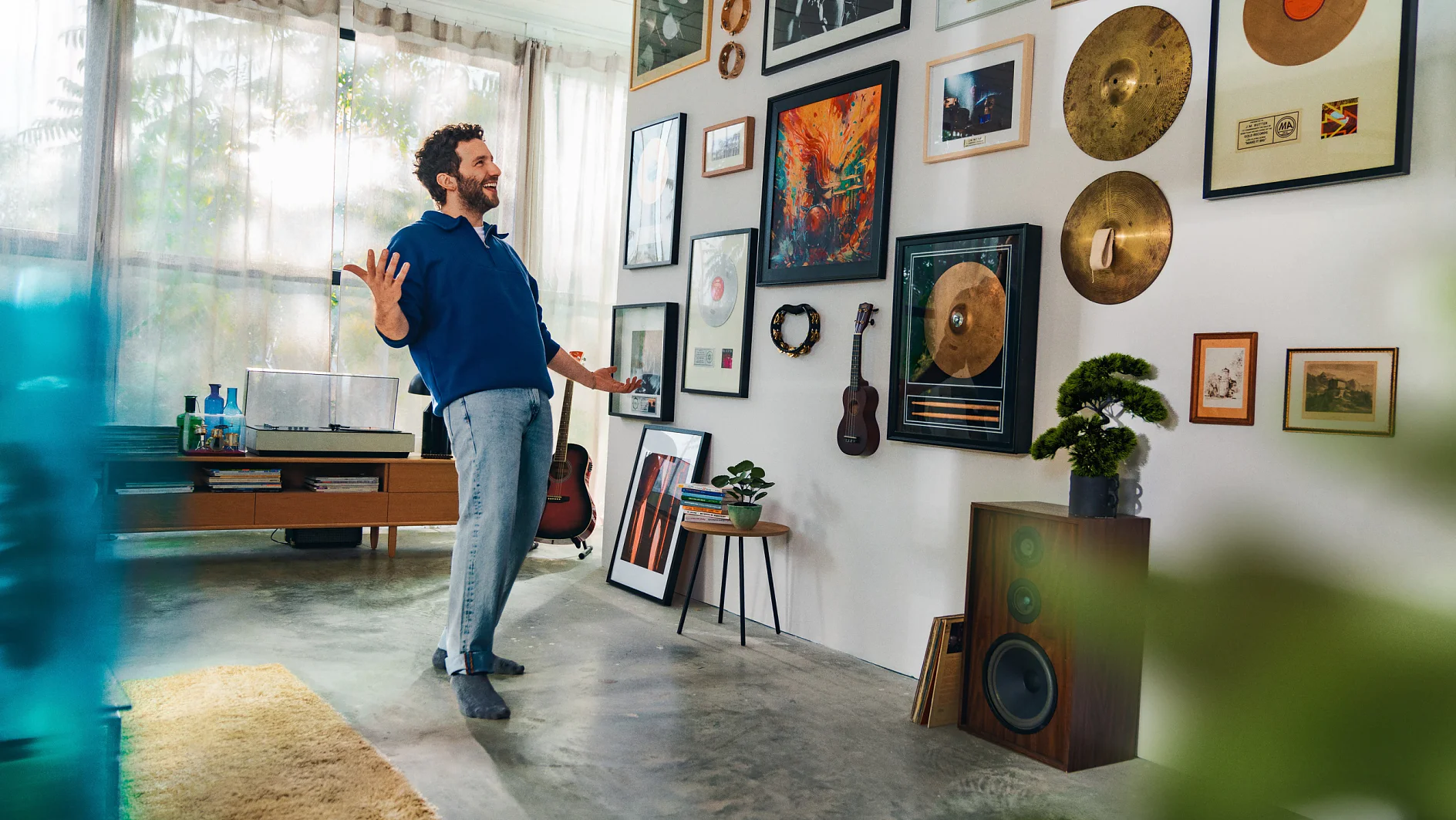 A man stands in a room looking at a wall adorned with various framed photos, art, and gold records. Theres a guitar and a small potted plant beside him. A record player and speakers are in the background, held together seamlessly with tesa tape, while sheer curtains let in light. (This text has been generated by AI)