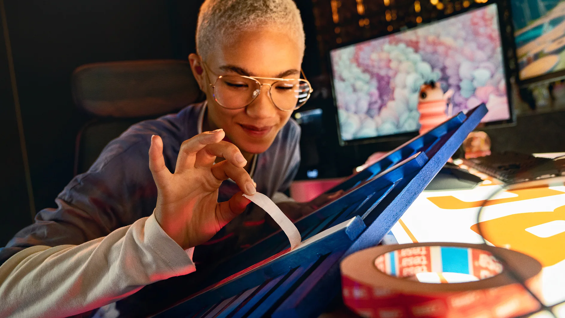 A person with short hair and glasses is seated at a desk, peeling tesa tape from a blue object. In the background, a computer monitor displays colorful graphics. Rolls of tesa tape and papers are visible on the desk. (This text has been generated by AI)