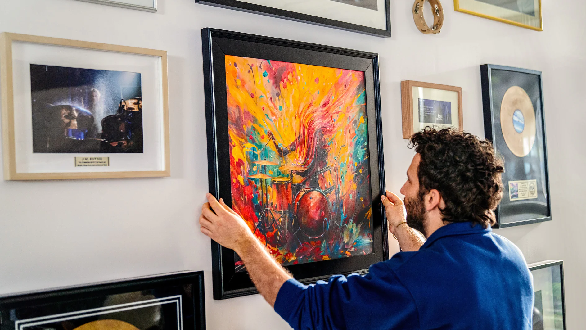 A man is hanging a colorful abstract painting on a wall filled with framed pictures and awards using tesa tape. He is wearing a blue shirt and focused on positioning the artwork, suggesting an appreciation for art and achievement. (This text has been generated by AI)