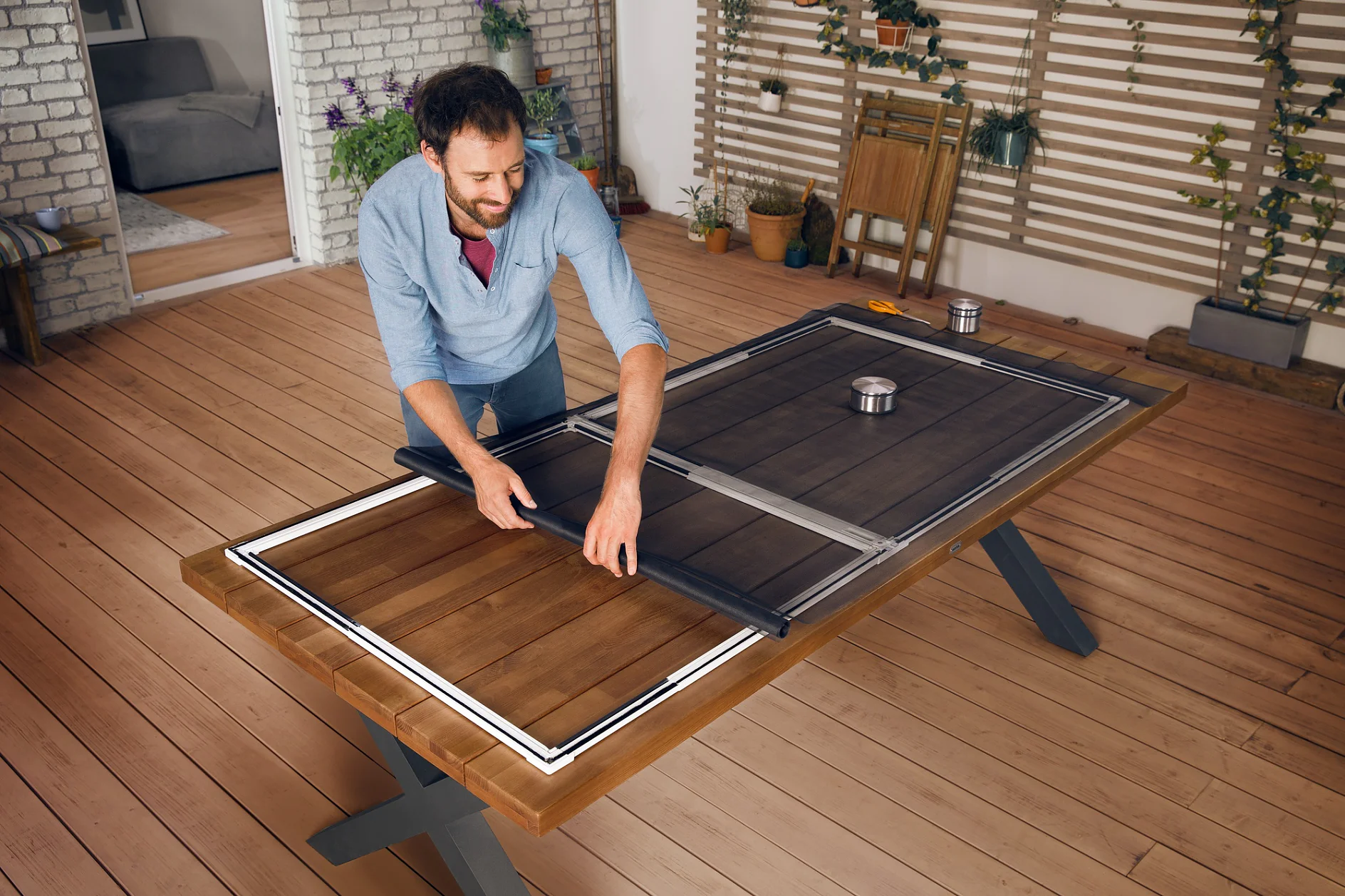 FALT Tür A person in a blue shirt assembles a table with a convertible table tennis surface in a room with wooden flooring and plant-covered walls using tesa tape. A chair and a plant are in the background. (This text has been generated by AI)