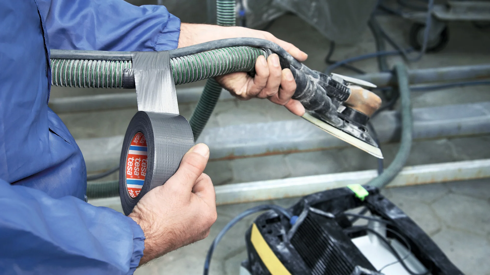 A person in a blue jacket is using silver tesa tape to secure a green ribbed hose to a metal tool with a rectangular base. The tool is connected to a piece of equipment with wheels in the background. (This text has been generated by AI)
