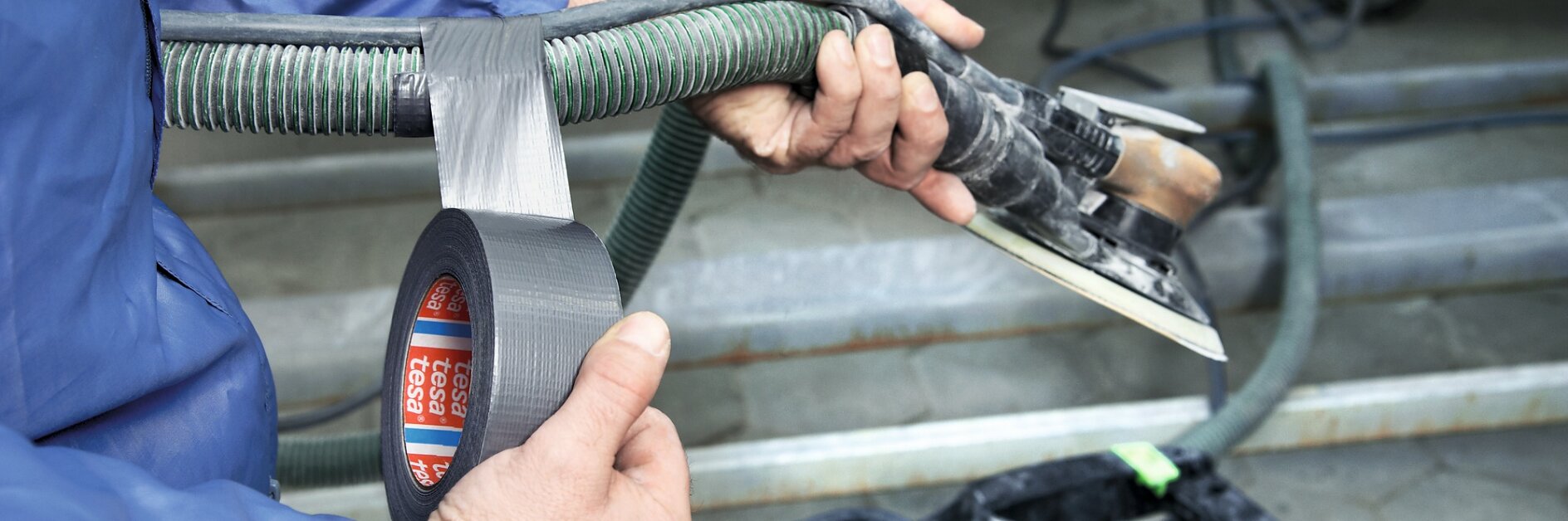 A person in a blue jacket is using silver tesa tape to secure a green ribbed hose to a metal tool with a rectangular base. The tool is connected to a piece of equipment with wheels in the background.