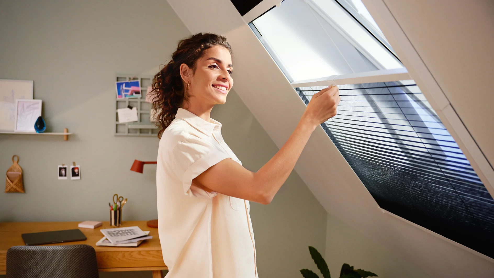 A woman in a light shirt adjusts the blinds on a skylight window in a modern home office. The room features a desk with papers, a lamp, and wall-mounted organizers. Soft natural light illuminates the space. (This text has been generated by AI)