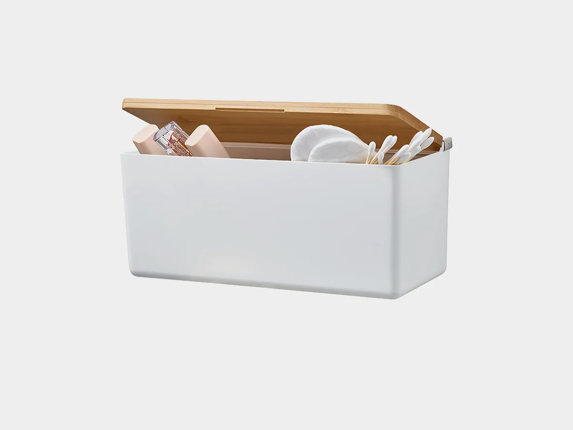 A rectangular white storage box with an open wooden lid contains various items, including cotton pads, tesa tape, and small bottles, against a plain white background. (This text has been generated by AI)