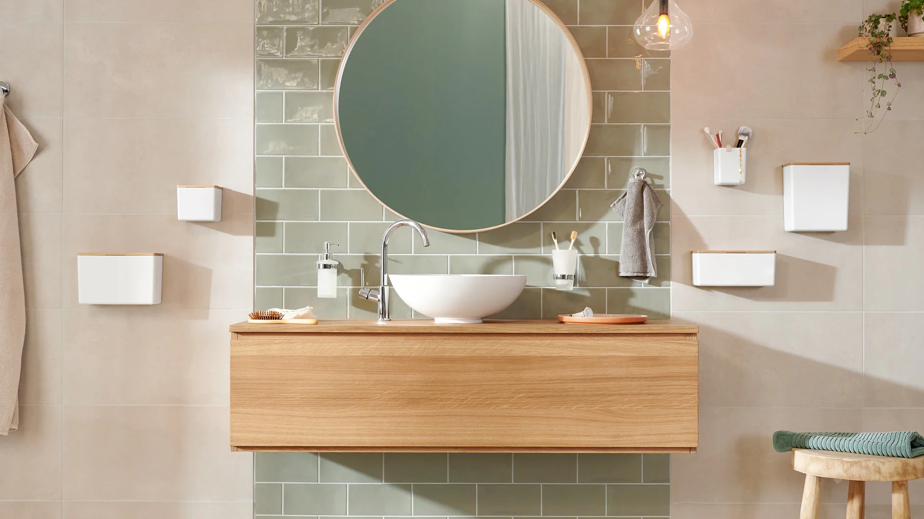 Modern bathroom with a wooden vanity and round mirror above. A white sink and silver faucet are centered on the vanity. Green tiled wall, pendant light, and assorted toiletries are visible. Neutral-toned walls and floor create a calming atmosphere, showcasing the seamless application of tesa tape for all mounting needs. (This text has been generated by AI)