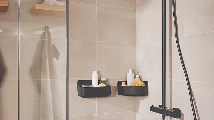 A modern shower with light beige tiled walls features black corner shelves holding toiletries and a shower scrub. A glass screen separates the shower area, and two towels hang on metal hooks secured with tesa tape. A black showerhead and controls are visible. (This text has been generated by AI)