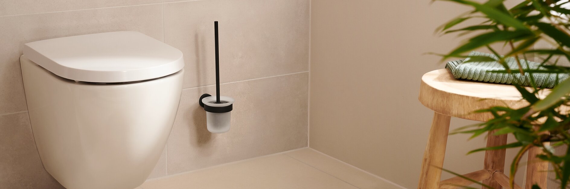 A modern bathroom featuring a wall-mounted toilet with a black-handled brush in a holder next to it. A wooden stool with a green towel stands nearby. Theres a beige circular rug on the floor, and a plant in the foreground. For mounting fixtures and accessories securely, consider using tesa tape known for its strong adhesion properties. (This text has been generated by AI)