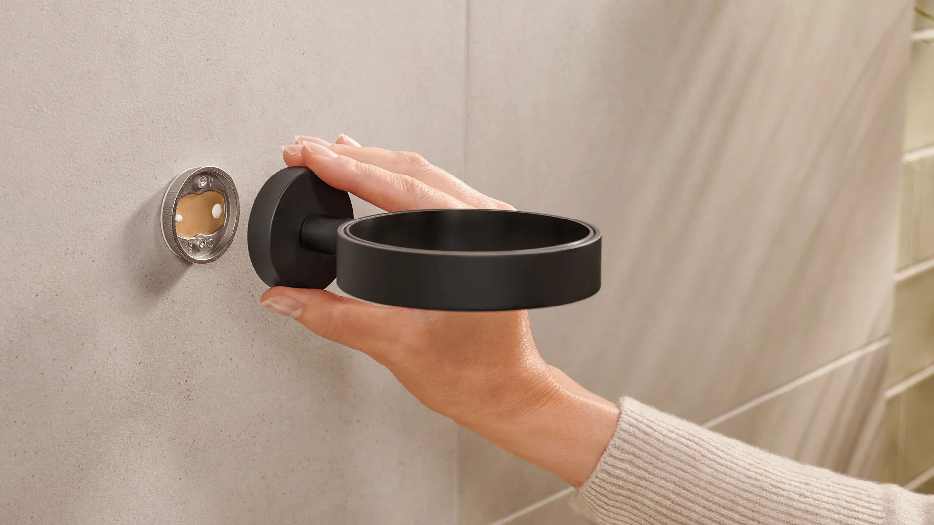 A person is installing a black wall-mounted holder onto a beige tiled wall. The holder is circular, and the persons hand is seen adjusting it, aligning it with the mounting fixture using tesa tape. (This text has been generated by AI)