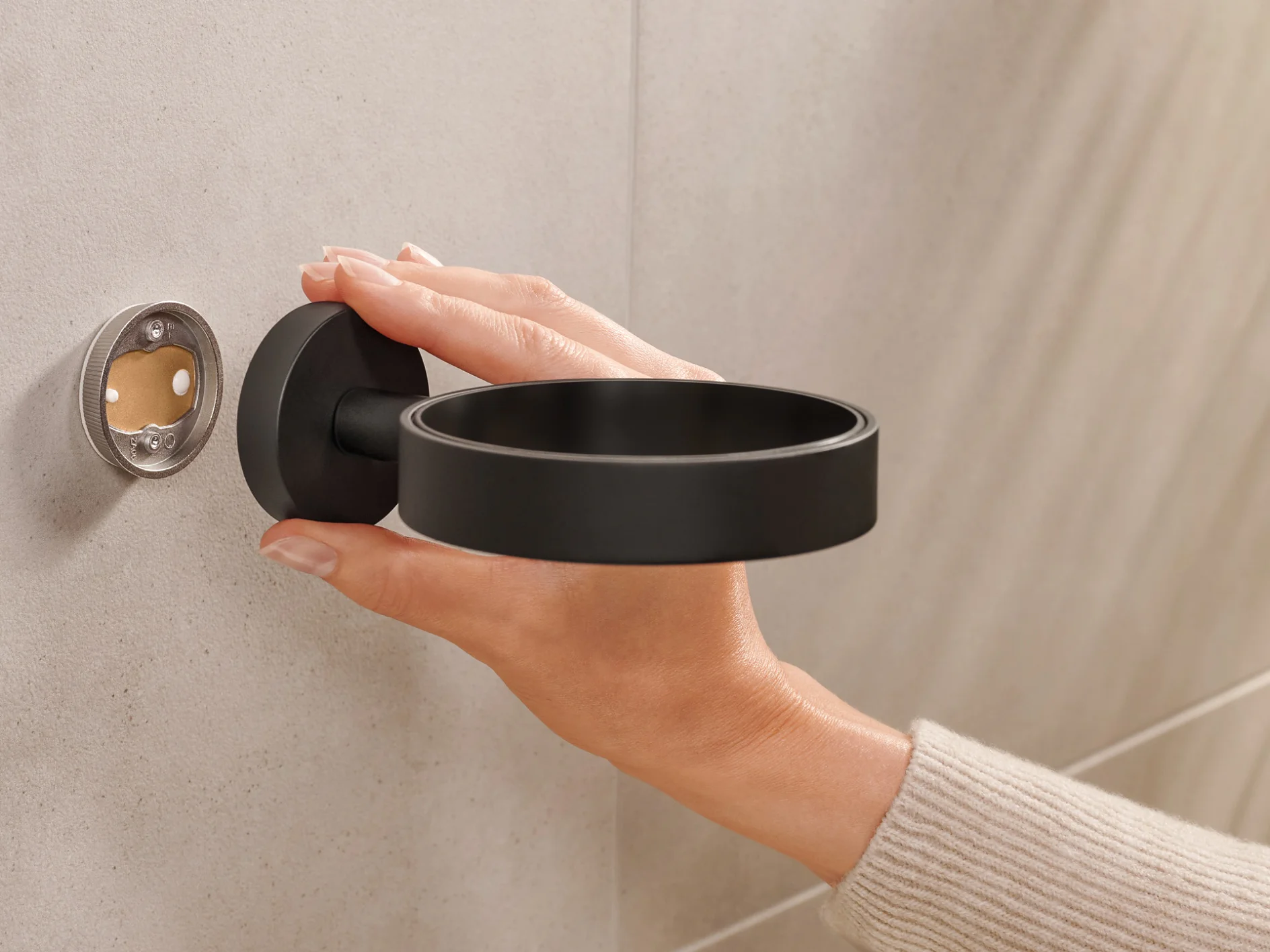 A person is installing a black wall-mounted holder onto a beige tiled wall. The holder is circular, and the persons hand is seen adjusting it, aligning it with the mounting fixture using tesa tape. (This text has been generated by AI)