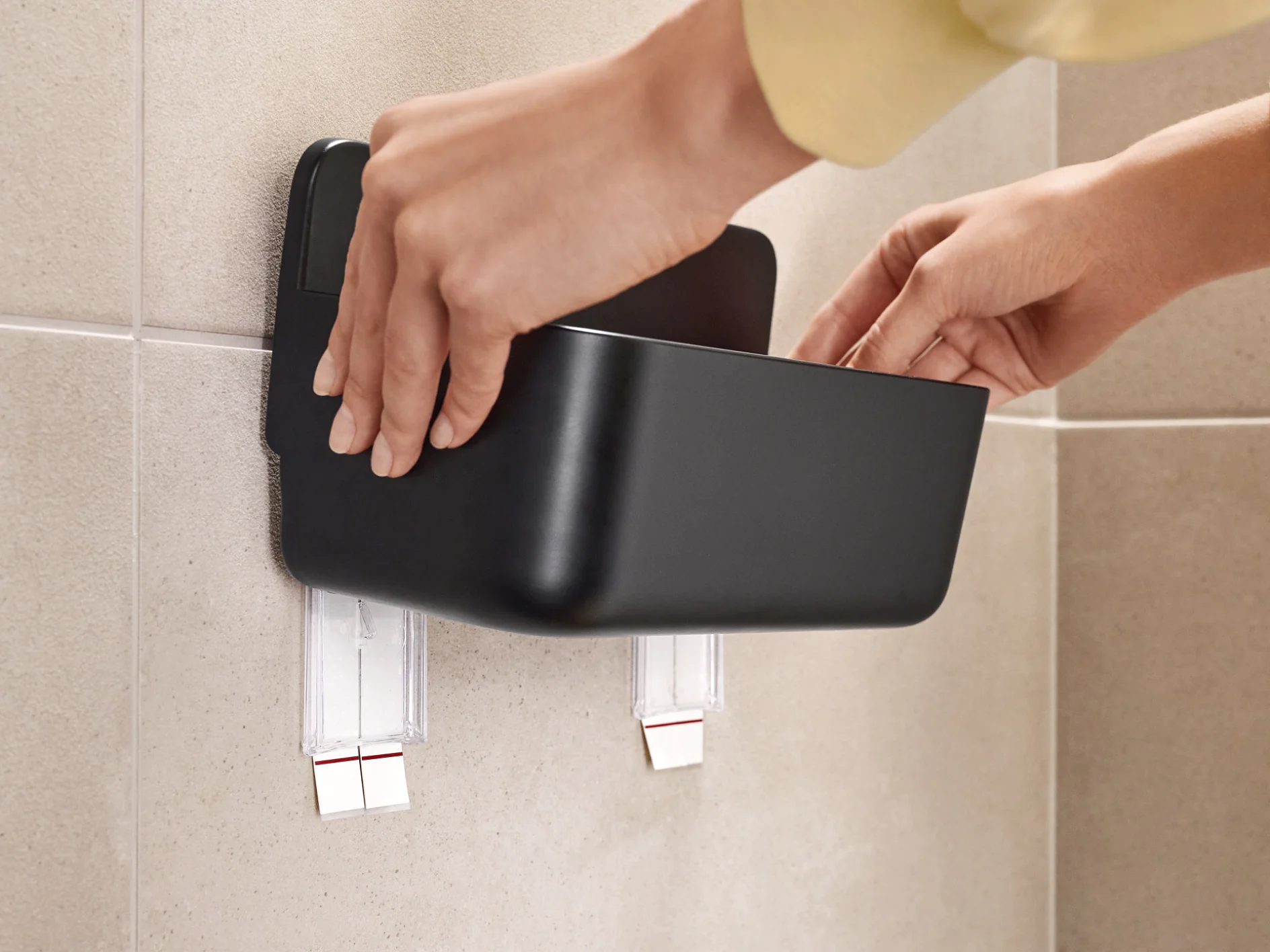 Hands are attaching a black container to a beige tiled wall using two strips of tesa tape. The container appears to be a wall-mounted organizer or storage shelf. (This text has been generated by AI)