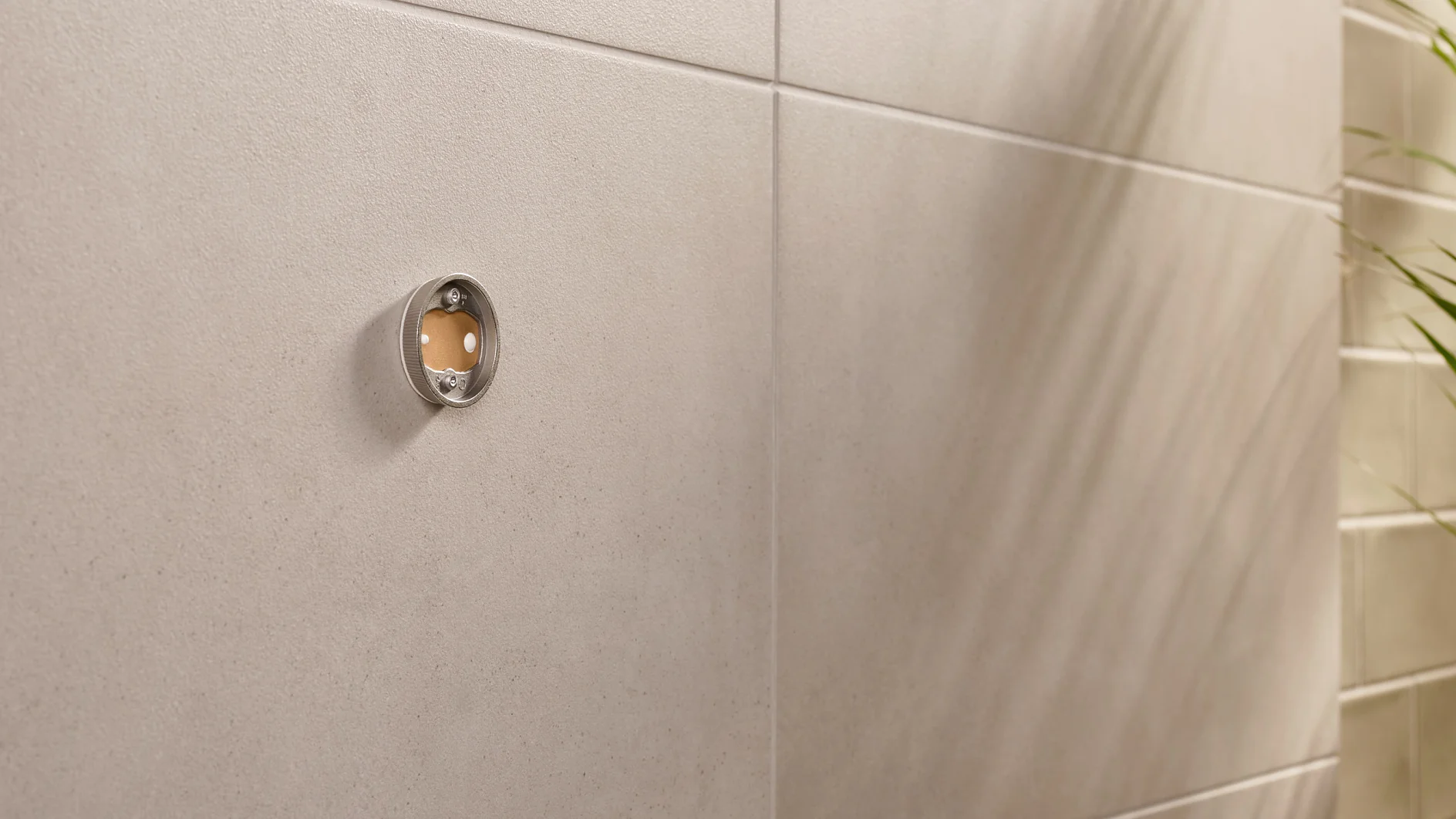 Close-up of a beige tiled wall with a circular metal fitting. The fitting has a transparent front, revealing a small reddish-brown circle inside. On the right, the shadow of a plant is visible on the wall. (This text has been generated by AI)