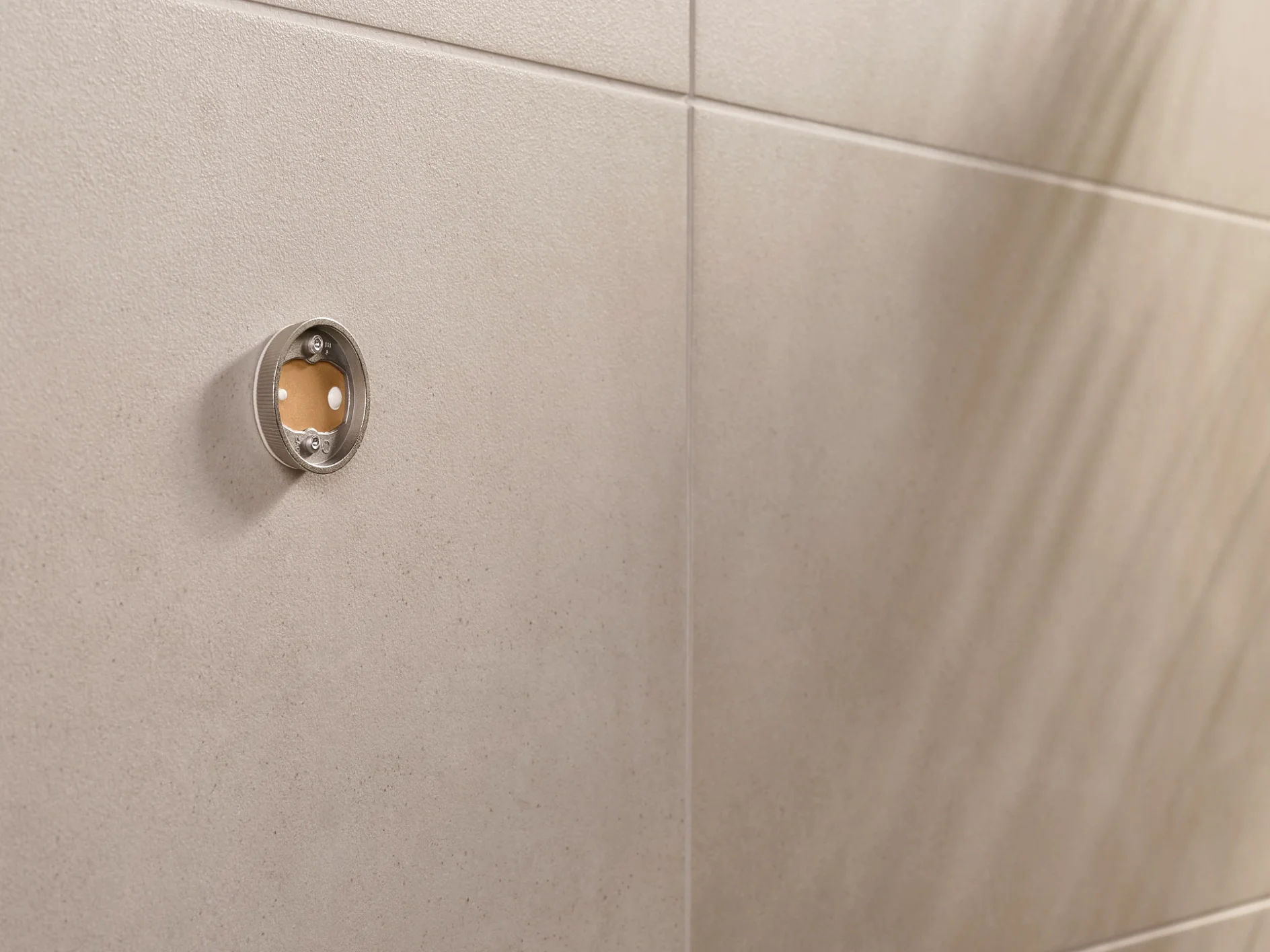 Close-up of a beige tiled wall with a circular metal fitting. The fitting has a transparent front, revealing a small reddish-brown circle inside. On the right, the shadow of a plant is visible on the wall. (This text has been generated by AI)