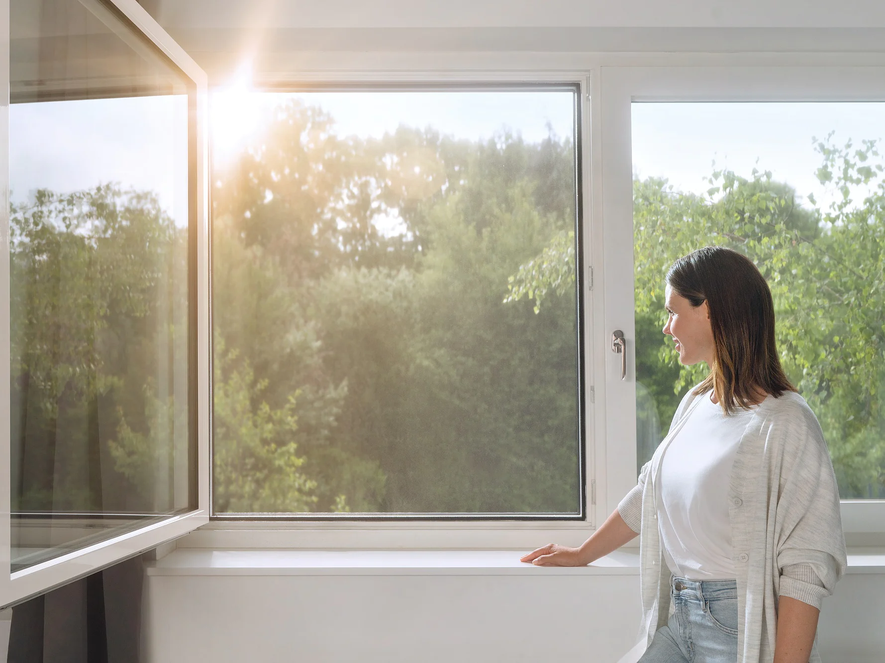 A woman stands by an open window, looking outside. Sunlight filters through, illuminating the greenery of trees in the background. She is wearing a light cardigan and jeans, with one hand resting on the windowsill secured with tesa tape. (This text has been generated by AI)