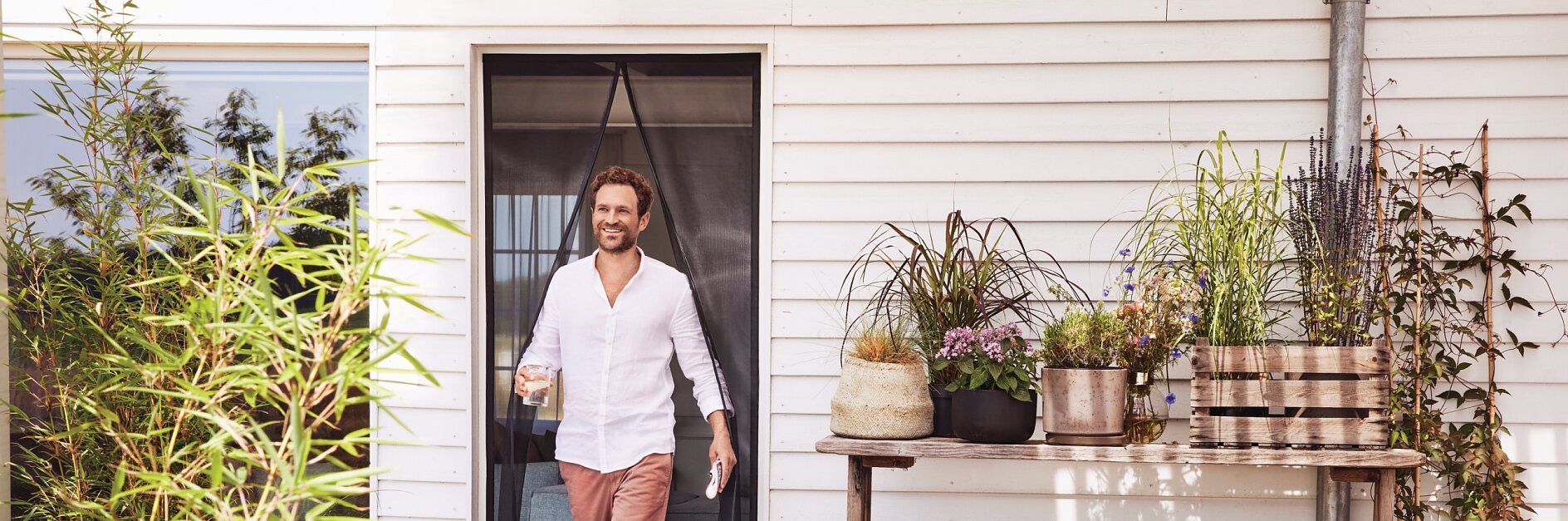 A man in a white shirt and pink pants holding a glass stands in the doorway of a white wooden house. The deck is adorned with potted plants and stacked firewood next to a table with more plants. (This text has been generated by AI)