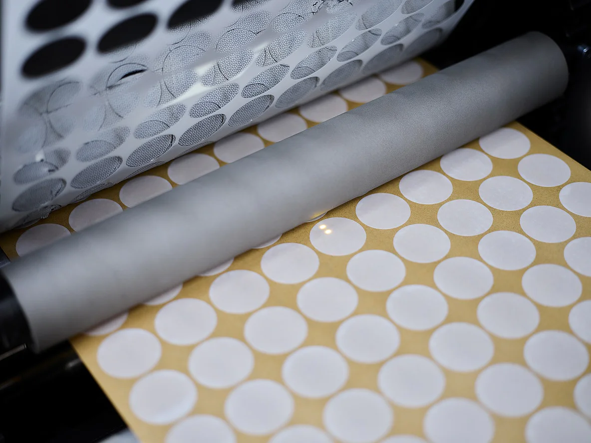 Close-up of a machine rolling a sheet of yellow paper with rows of white circular stickers made from tesa tape, with additional white tesa tape stickers visible on the upper part of the machine.
