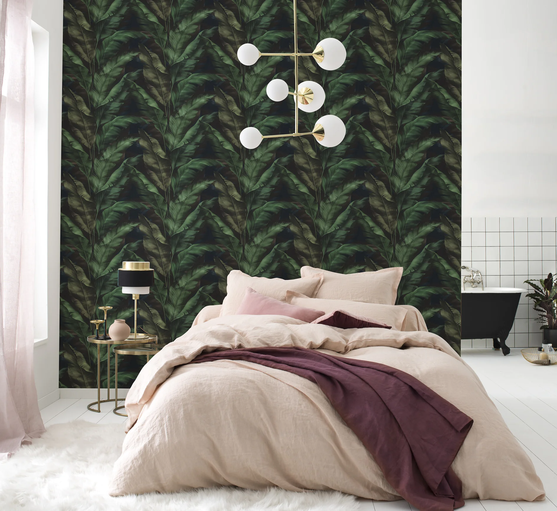 A modern bedroom with a bed covered in beige and mauve bedding, a gold light fixture, a side table with a lamp, and a feature wall with a dark green leafy pattern created using tesa tape. A black bathtub is visible in the background. (This text has been generated by AI)