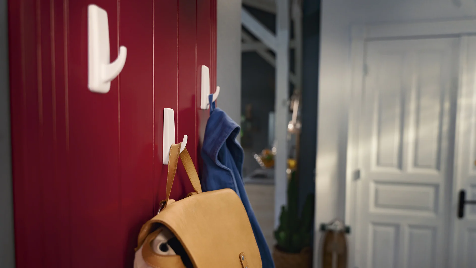 A red wooden wall with three white hooks. A tan leather bag hangs on the lower hook, secured by tesa tape, while a blue jacket hangs on the middle hook, also held up by tesa tape. A hallway and a white door are visible in the background. (This text has been generated by AI)