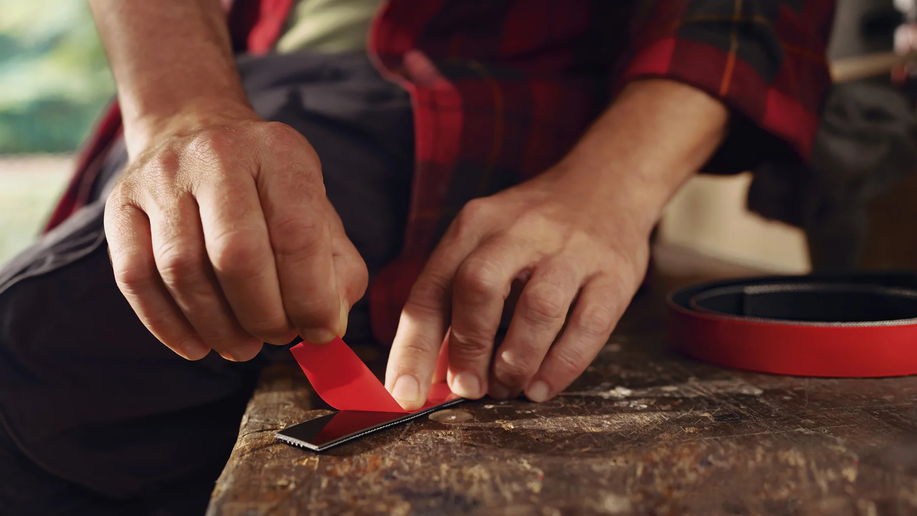 A person in a red and black plaid shirt is cutting red adhesive tesa tape on a wooden surface with a utility knife. A roll of the same tesa tape lies nearby. The persons hands are focused on the task, and the background is slightly blurred. (This text has been generated by AI)