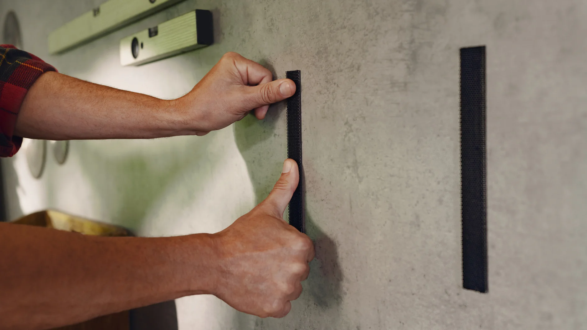 A person is applying two vertical strips of tesa tape to a wall. They press one strip with their thumb while holding the other strip nearby. A level tool is seen in the background on the wall. (This text has been generated by AI)