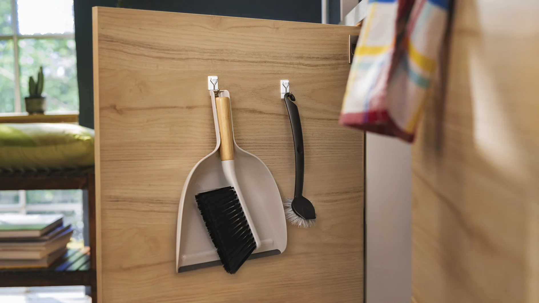 A wooden cabinet door features two hooks holding a dustpan with a brush and a dishwashing brush secured with tesa tape. A striped cloth is partially visible on the right side. In the background, theres a window and a cushioned bench with books. (This text has been generated by AI)