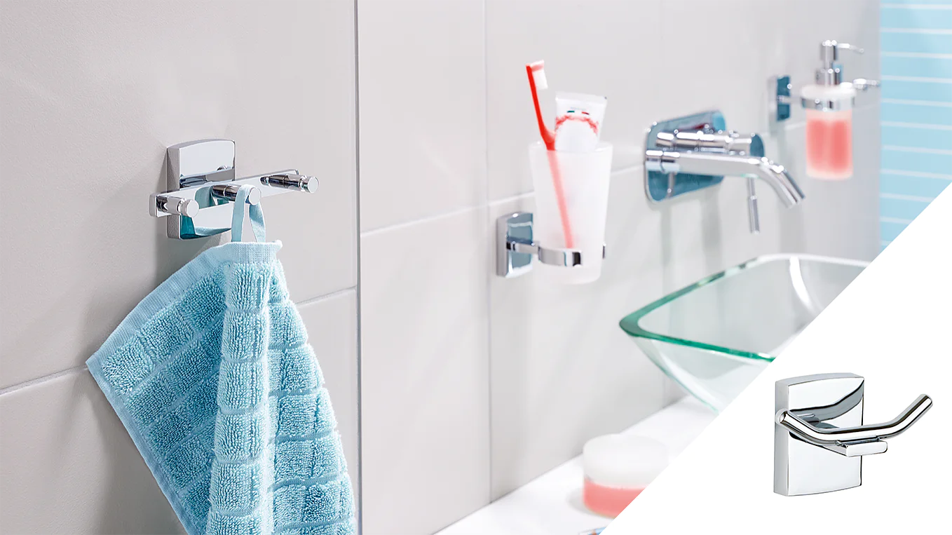 A modern bathroom setup with a light blue towel hanging on a chrome hook using tesa tape, a clear glass holding a red toothbrush above, and a soap dispenser on the right. The image also features a smaller inset of the chrome hook. (This text has been generated by AI)